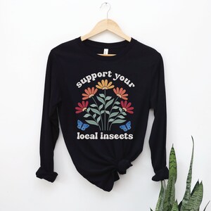 A black unisex long sleeve crew neck t-shirt with the words "support your local insects" in white text around an image of colorful flowers and butterflies