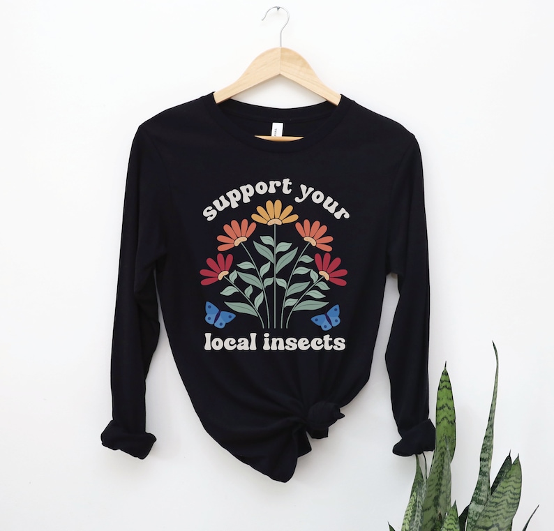 A black unisex long sleeve crew neck t-shirt with the words "support your local insects" in white text around an image of colorful flowers and butterflies