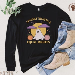 May include: A black long-sleeve shirt featuring a graphic of a white ghost, two pumpkins, and a yellow moon with purple clouds. The text reads "SPOOKY NIGHTS & EQUAL RIGHTS" in yellow. A small circular logo in the corner shows a raised fist icon.