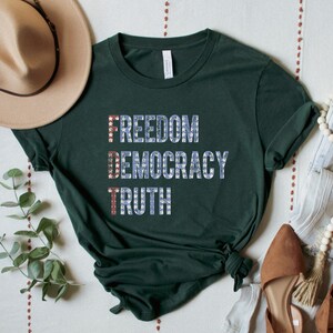 A forest green unisex crew neck short sleeve Bella+Canvas t-shirt that says "FREEDOM DEMOCRACY TRUTH" on the chest in three lines in blue block text with white stars inside the letters. The first letter of each word in in red with white stars, subtly spelling "FDT" down the shirt