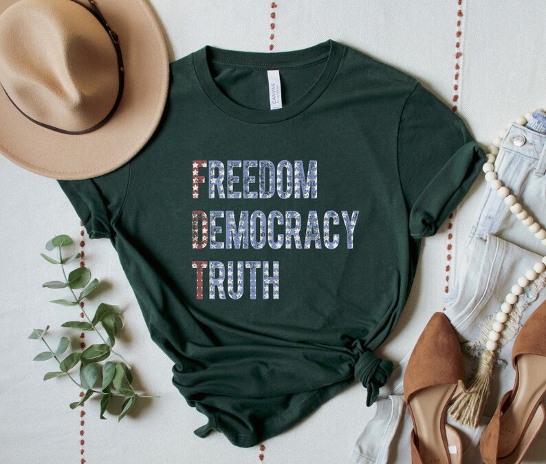 A forest green unisex crew neck short sleeve Bella+Canvas t-shirt that says "FREEDOM DEMOCRACY TRUTH" on the chest in three lines in blue block text with white stars inside the letters. The first letter of each word in in red with white stars, subtly spelling "FDT" down the shirt