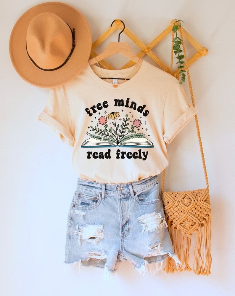 A natural (light beige) unisex crew neck short sleeve t-shirt with black text saying "free minds read freely" around a graphic of a book with colorful flowers and stars coming out of it