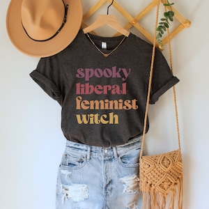 A dark grey heather unisex crew neck t-shirt with the phrase "spooky liberal feminist witch" in purple, red, orange, and yellow text