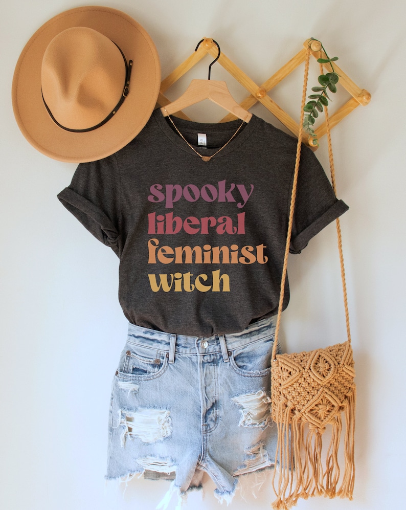 A dark grey heather unisex crew neck t-shirt with the phrase "spooky liberal feminist witch" in purple, red, orange, and yellow text