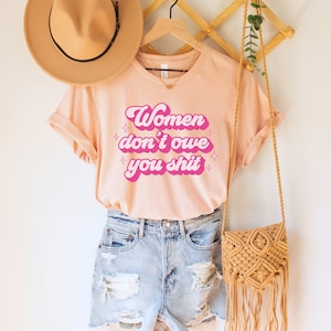 A heather peach unisex crew neck t-shirt with the words "women don't owe you shit" in retro pink text with retro pink stars