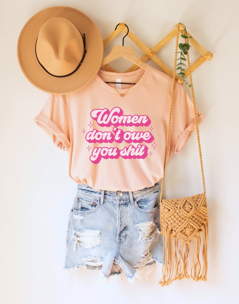 A heather peach unisex crew neck t-shirt with the words "women don't owe you shit" in retro pink text with retro pink stars