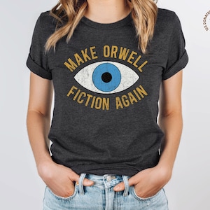 A dark grey heather unisex crew neck Bella Canvas brand t-shirt that says "MAKE ORWELL FICTION AGAIN" in orange text with a black outline, around an image of a blue eye. The graphic has a distressed vintaged look.