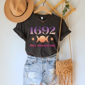 Puede incluir: Una camiseta gris oscuro con un gr&aacute;fico rosa y morado del n&uacute;mero 1692, tres lunas y estrellas. El texto "they missed one" est&aacute; impreso debajo del gr&aacute;fico.
