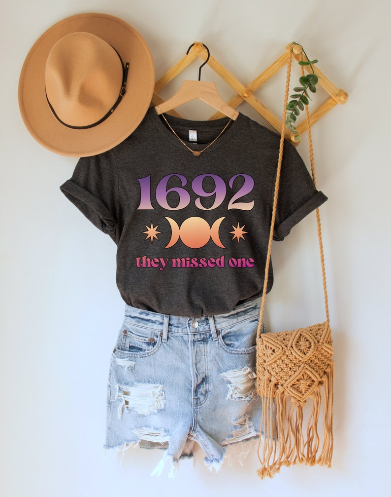 Puede incluir: Una camiseta gris oscuro con un gr&aacute;fico rosa y morado del n&uacute;mero 1692, tres lunas y estrellas. El texto "they missed one" est&aacute; impreso debajo del gr&aacute;fico.
