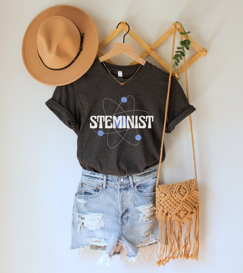 May include: A dark gray t-shirt with a white atom graphic and the text "STEMINIST" in white letters. The shirt is being displayed on a wooden hanger with a brown hat and a brown crocheted bag.