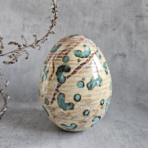 Op de afbeelding: Een keramisch ei met een beige achtergrond en een patroon van bruine lijnen en turquoise vlekken en swirls.