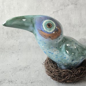 Op de afbeelding: Een keramische vogelsculptuur in blauw- en groentinten, zittend op een geweven nest. De vogel heeft een lange, gebogen snavel en een gedetailleerd oog met bruine en gouden accenten. Het beeld is een decoratief item.