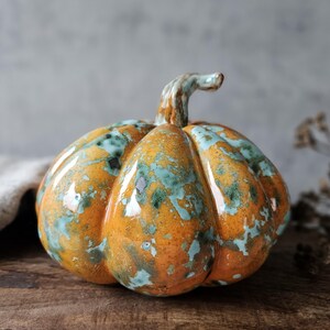 Op de afbeelding: Een keramieken pompoensculptuur met een glanzende afwerking en een gevlekt patroon van levendig oranje en gedempt blauwgroen. De pompoen heeft een rond, geribbeld lichaam en een gebogen steeltje aan de bovenkant met een glad oppervlak.