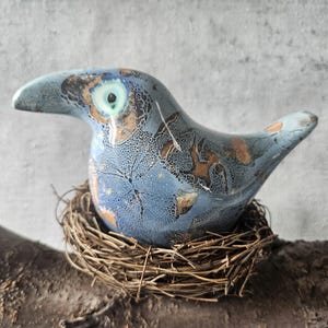 Może przedstawiać: Ceramiczna figurka ptaka z niebiesko-brązową, nakrapianą glazurą, siedząca w wiklinowym gnieździe. Ptak ma długi dziób i jedno oko. Gniazdo wykonane jest z gałązek i spoczywa na drewnianej gałęzi.