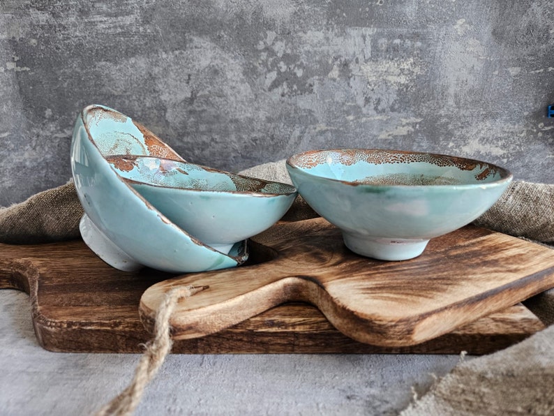 K&ouml;nnte beinhalten: Zwei tealblaue Keramikschalen mit einem gesprenkelten &Uuml;berzug, die auf einem Holzbrett liegen. Die Schalen sind &uuml;bereinander gestapelt.