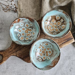 K&ouml;nnte beinhalten: Drei kleine Keramikschalen mit hellblauer Glasur und braunem, gesprenkeltem Muster. Die Schalen sind auf einem Holztablett angeordnet.