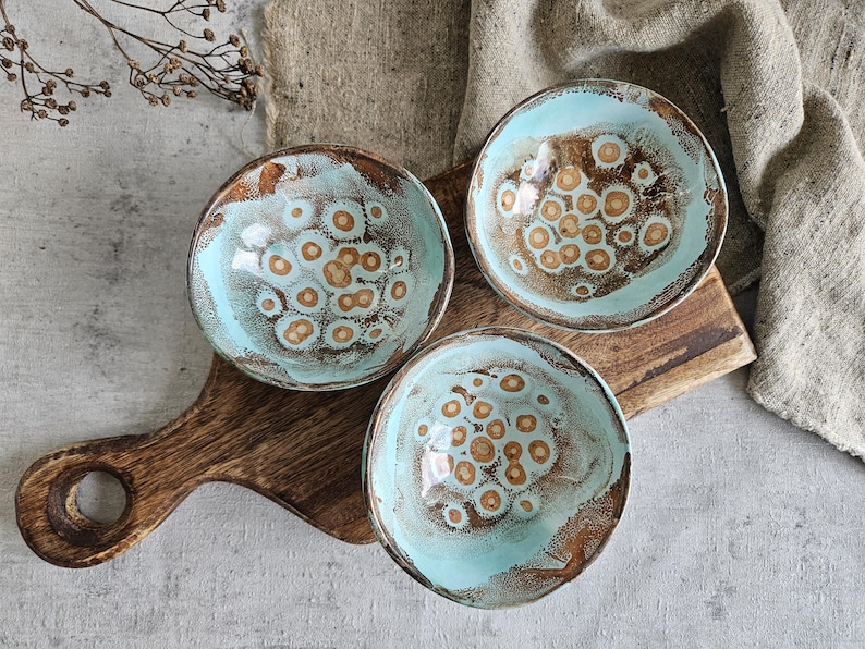 K&ouml;nnte beinhalten: Drei kleine Keramikschalen mit hellblauer Glasur und braunem, gesprenkeltem Muster. Die Schalen sind auf einem Holztablett angeordnet.