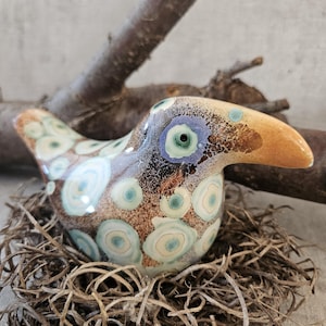 Peut inclure: Figurine d'oiseau en céramique avec un motif tacheté marron, bleu et crème, nichée dans un nid d'herbe sèche. L'oiseau a un long bec orange et se détache sur un fond neutre avec une branche d'arbre.