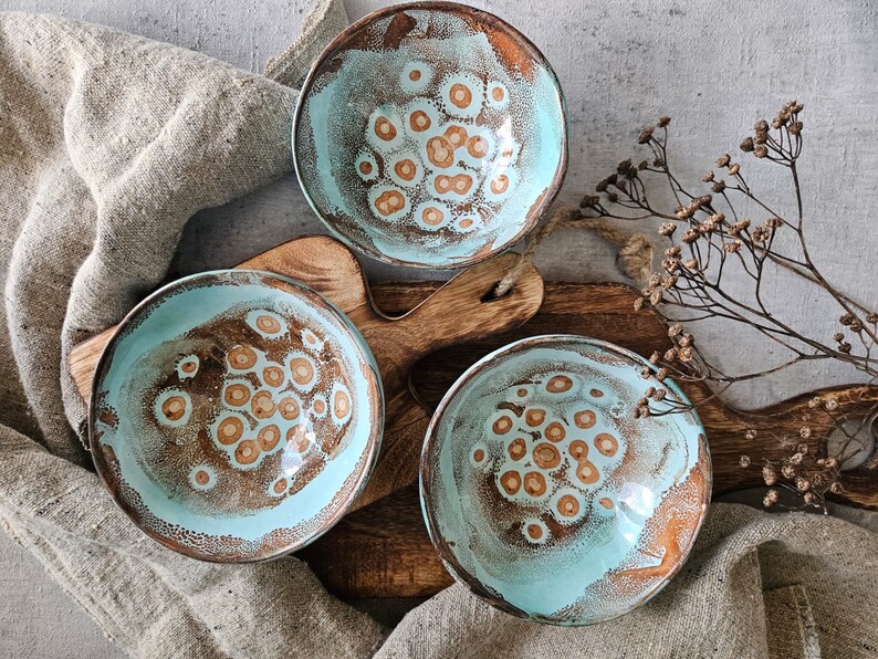 K&ouml;nnte beinhalten: Drei tealblaue Keramikschalen mit braunem, gesprenkeltem Design. Die Schalen sind auf einer Holzoberfl&auml;che angeordnet.