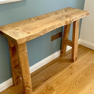 May include: A wooden console table with a live edge top. The table is supported by two A-frame legs. The wood has a warm, light brown colour and visible grain. The table is approximately 152 cm long.