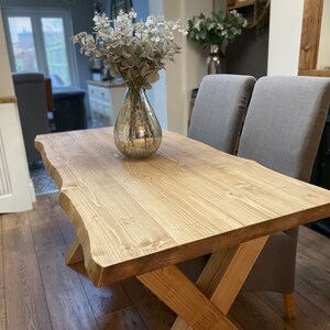 May include: A rustic wooden dining table with a natural edge and an X-shaped base. The table is made of light-colored wood and is paired with two gray upholstered chairs. A decorative vase with silver foliage sits on the table.