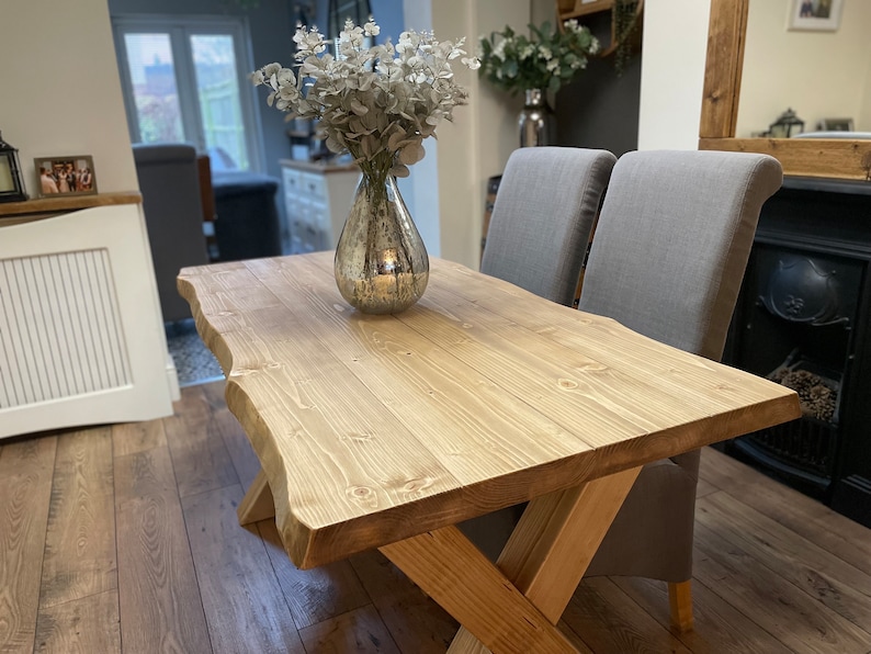 May include: A rustic wooden dining table with a natural edge and an X-shaped base. The table is made of light-colored wood and is paired with two gray upholstered chairs. A decorative vase with silver foliage sits on the table.