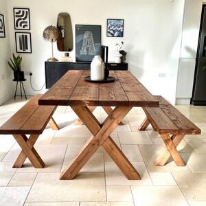 May include: A rustic wooden dining table with two matching benches. The table has a dark brown stain and a cross-shaped base. The benches are also stained dark brown and have a simple, straight design.
