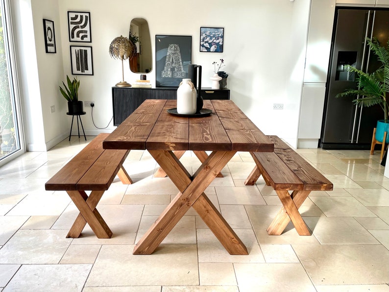 May include: A rustic wooden dining table with two matching benches. The table has a dark brown stain and a cross-shaped base. The benches are also stained dark brown and have a simple, straight design.