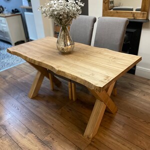 May include: A natural wood dining table with a live edge design. The table has an X-shaped base and is approximately 60 inches long. A silver vase with white flowers sits on the table. Two gray chairs are behind the table.