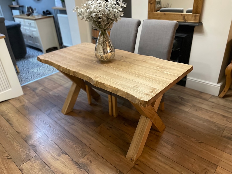 May include: A natural wood dining table with a live edge design. The table has an X-shaped base and is approximately 60 inches long. A silver vase with white flowers sits on the table. Two gray chairs are behind the table.