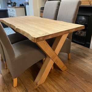 May include: A light-colored wooden dining table with a natural edge, supported by an X-shaped base. Two gray upholstered chairs are positioned on either side. The table is approximately 60 inches long.