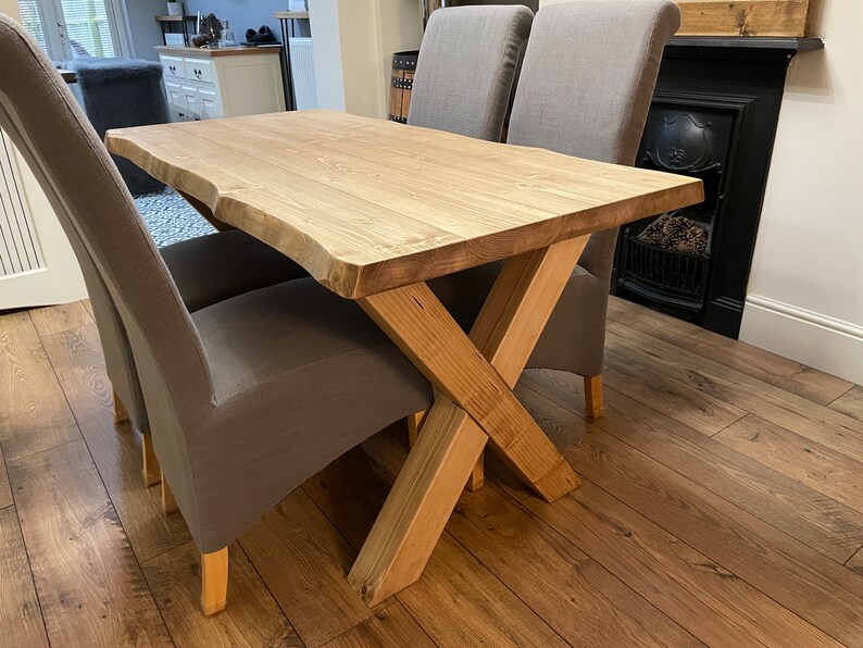 May include: A light-colored wooden dining table with a natural edge, supported by an X-shaped base. Two gray upholstered chairs are positioned on either side. The table is approximately 60 inches long.