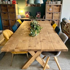May include: A rustic wooden dining table with a cross-shaped base. The table is made of natural wood with a light brown finish. It is set with a centerpiece of greenery and has six chairs around it, two of which are yellow velvet and two are gray.