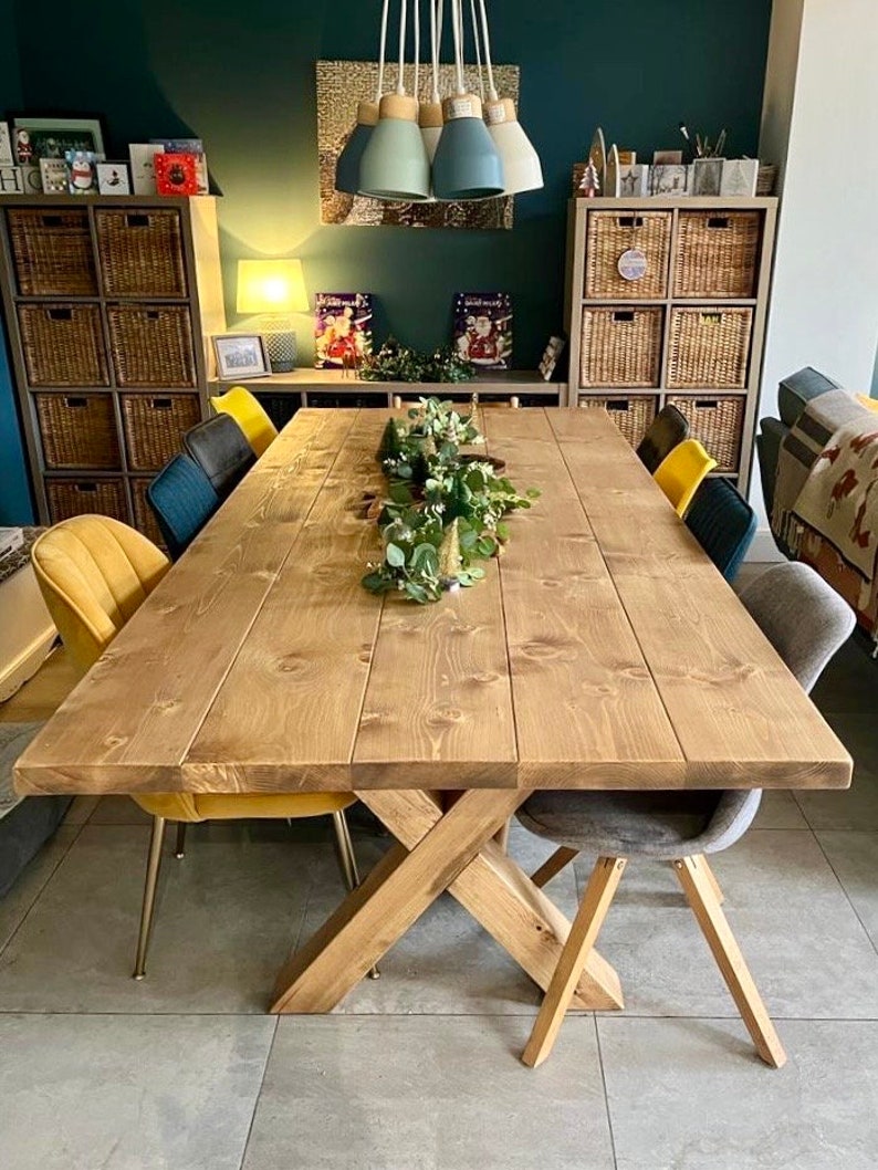 May include: A rustic wooden dining table with a cross-shaped base. The table is made of natural wood with a light brown finish. It is set with a centerpiece of greenery and has six chairs around it, two of which are yellow velvet and two are gray.