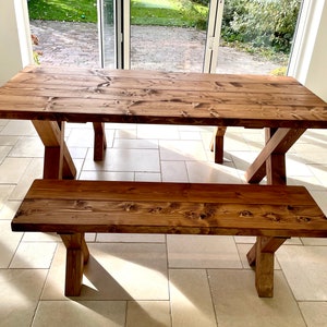 May include: A rustic wooden dining table and bench set with a natural brown finish. The table has a rectangular top and a simple X-shaped base. The bench has a similar design with a shorter top and matching base.