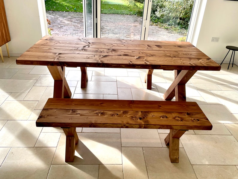 May include: A rustic wooden dining table and bench set with a natural brown finish. The table has a rectangular top and a simple X-shaped base. The bench has a similar design with a shorter top and matching base.