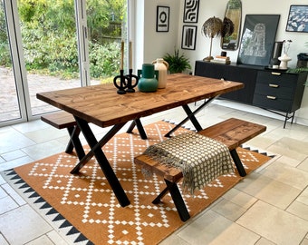 May include: A wooden dining table with a dark finish and black metal cross legs. Two matching benches and a patterned rug are beneath the table. Decorative vases and candles are on the table. The room has large windows.