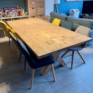 May include: A large wooden dining table with a natural finish. The table has a cross-shaped base and is surrounded by chairs with different colored cushions.