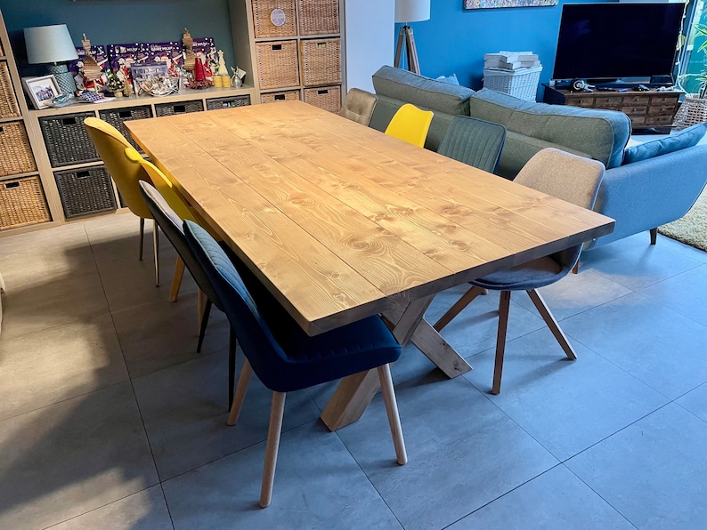 May include: A large wooden dining table with a natural finish. The table has a cross-shaped base and is surrounded by chairs with different colored cushions.