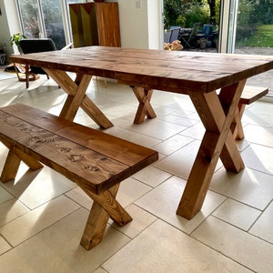 May include: A rustic wooden dining table and bench set with a natural brown finish. The table has a rectangular top and X-shaped legs. The bench has a rectangular top and X-shaped legs. The set is made of solid wood and has a natural, rustic look.