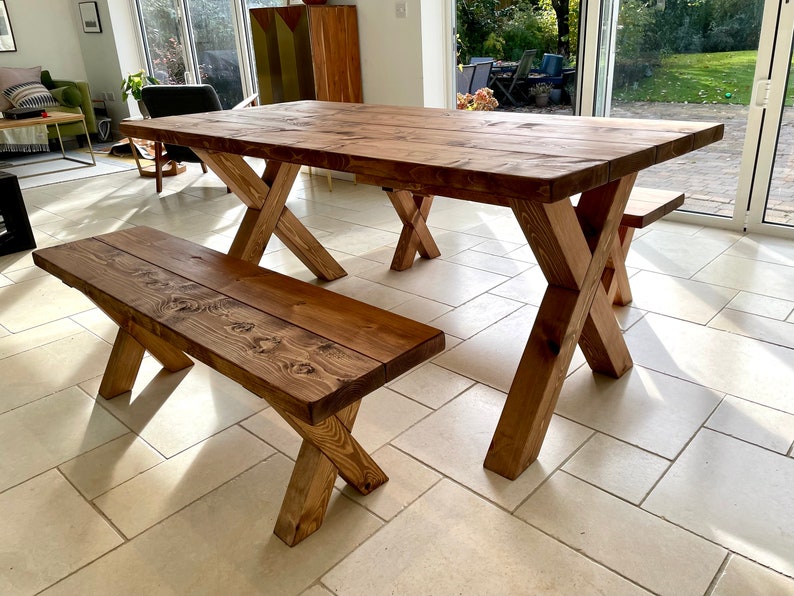 May include: A rustic wooden dining table and bench set with a natural brown finish. The table has a rectangular top and X-shaped legs. The bench has a rectangular top and X-shaped legs. The set is made of solid wood and has a natural, rustic look.