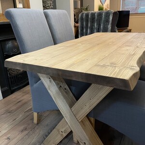 May include: A light-colored wooden dining table with a natural edge and an X-shaped base. The table is paired with two gray upholstered chairs. The table's dimensions are approximately 60 inches long and 30 inches wide.