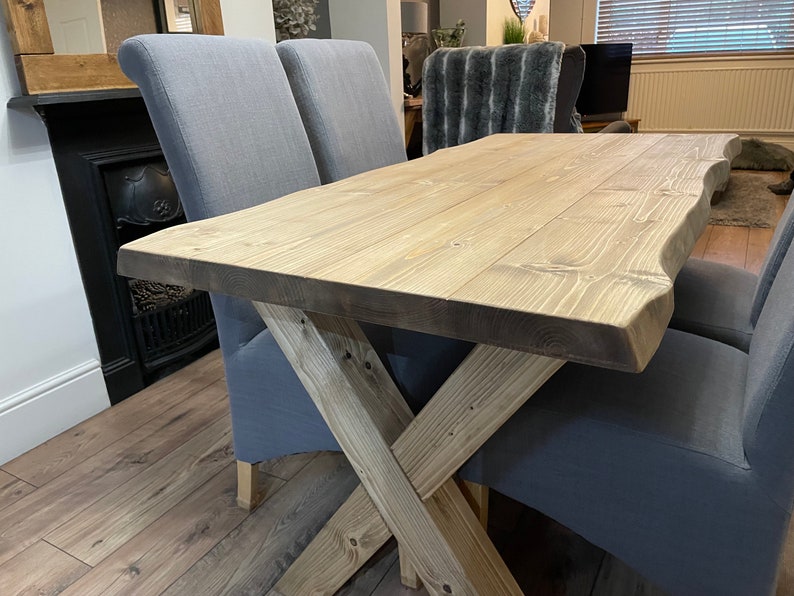 May include: A light-colored wooden dining table with a natural edge and an X-shaped base. The table is paired with two gray upholstered chairs. The table's dimensions are approximately 60 inches long and 30 inches wide.