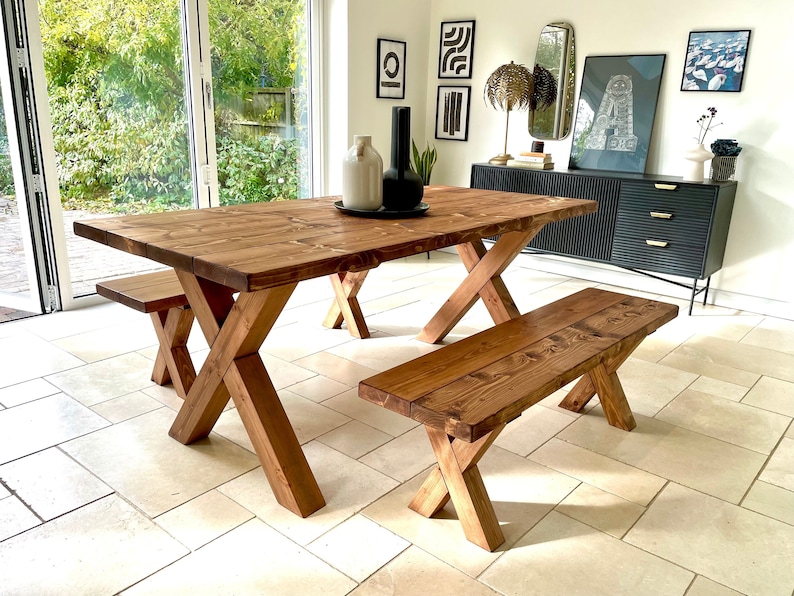 May include: A rustic wooden dining table and two matching benches. The table has a natural wood finish and a cross-shaped base. The benches are also made of wood and have a similar cross-shaped base.