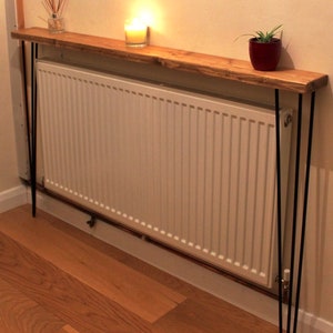 May include: A rustic wooden shelf with black metal hairpin legs sits above a white radiator. The shelf is decorated with a candle and a potted plant.