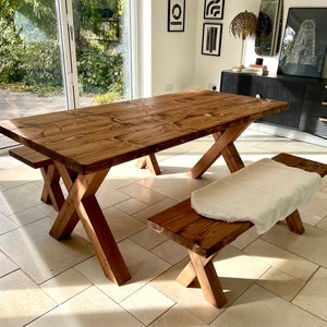 May include: A rustic wooden dining table with a trestle base and a matching bench. The table and bench are made of natural wood with a warm brown finish. The table is set in a room with a large window and a tiled floor.