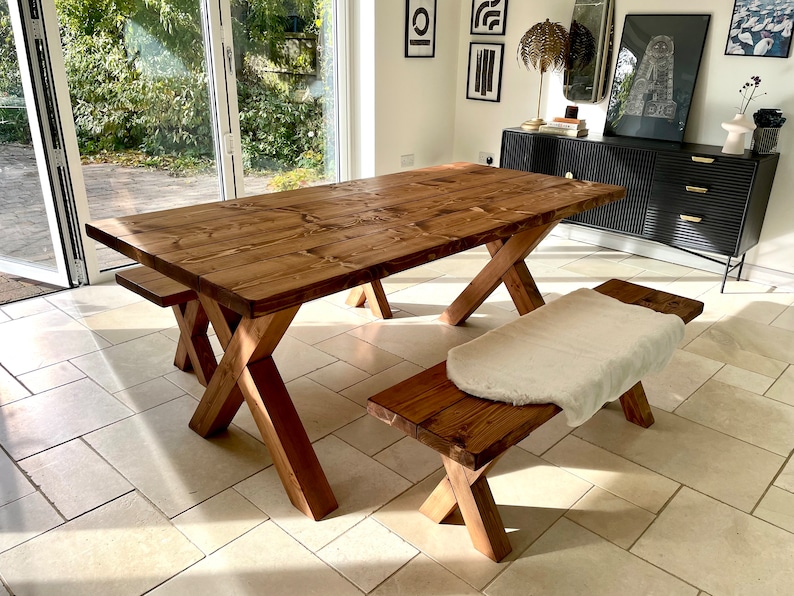 May include: A rustic wooden dining table with a trestle base and a matching bench. The table and bench are made of natural wood with a warm brown finish. The table is set in a room with a large window and a tiled floor.