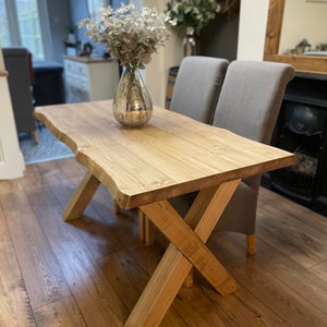 May include: A light-colored wooden dining table with a natural edge and an X-shaped base. The table is approximately 60 inches long. A vase with dried flowers sits on the table, with two gray upholstered chairs behind it.