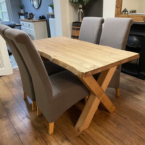 May include: A wooden dining table with a natural edge and a light finish, paired with four gray upholstered chairs. The table has a unique X-shaped base. The chairs have curved backs and wooden legs.