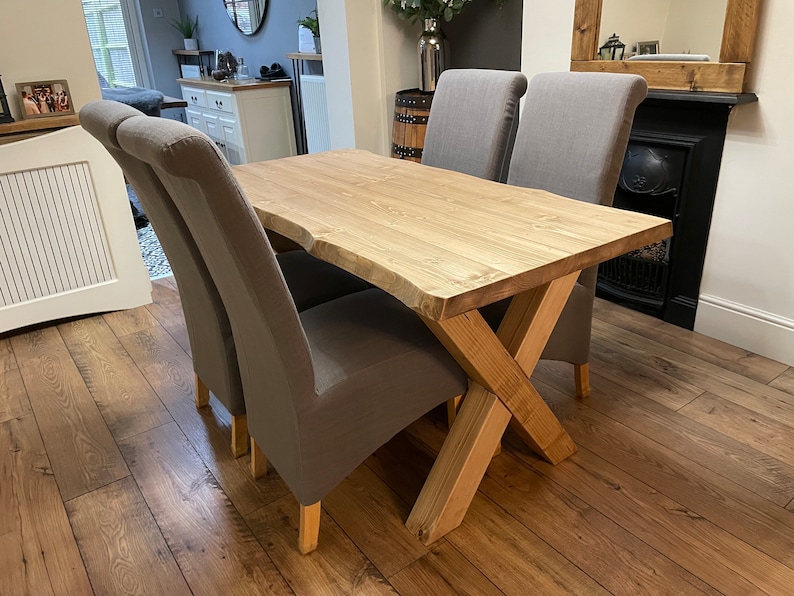 May include: A wooden dining table with a natural edge and a light finish, paired with four gray upholstered chairs. The table has a unique X-shaped base. The chairs have curved backs and wooden legs.